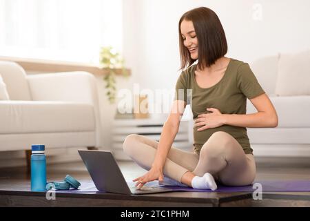 La giovane donna incinta pratica yoga prenatale usando il computer a casa Foto Stock