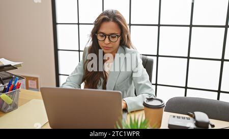 Seria ma rilassata, una giovane donna ispanica prende il ruolo di un capo elegante. una bella professionista, che lavora diligentemente sul suo computer portatile alla h Foto Stock