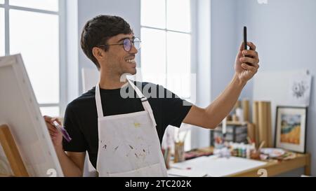 Un giovane artista ispanico sorridente che scatta una splendida foto di selfie con il suo smartphone nel vivace studio d'arte, tela in una mano, pennello in t Foto Stock