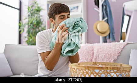 Bel giovane ispanico si rilassa tranquillamente sul divano, profumando allegramente il fresco profumo di vestiti piegati, godendosi le faccende domestiche al suo interno Foto Stock