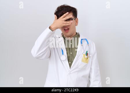 Giovane uomo non binario che indossa uniforme da medico e stetoscopio sbircia in shock coprendo viso e occhi con mano, guardando attraverso le dita con imbarazzo Foto Stock