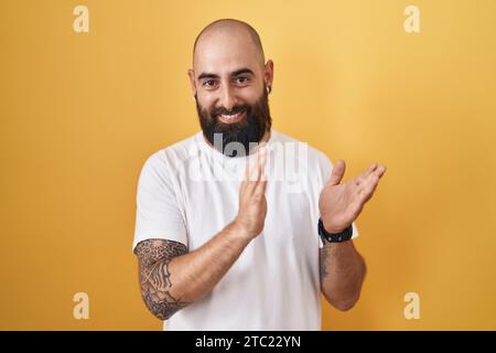 Giovane ispanico con barba e tatuaggi in piedi su sfondo giallo che applaude felici e gioiosi, sorridendo mani orgogliose insieme Foto Stock