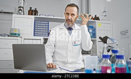 Uomo di mezza età dai capelli grigi ispanici, scienziato di laboratorio, impegnato nella ricerca scientifica, ascoltando un messaggio vocale sul suo smartphone in laboratorio Foto Stock