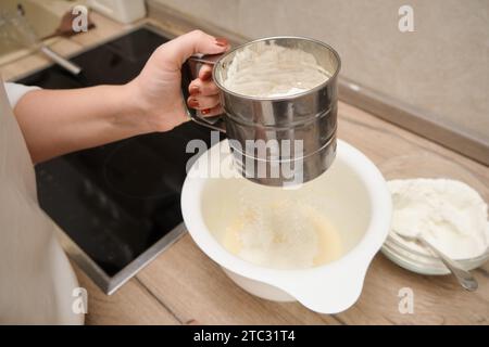 Le mani femminili versano la farina in una ciotola per la torta in cucina e setacciano la farina. Foto Stock