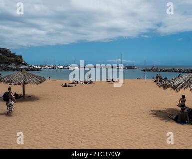 Machico, Madeira, Portogallo, 17 maggio 2022: Persone che si rilassano e nuotano nella spiaggia di sabbia dorata e nel porticciolo di Machico con barche da pesca e yacht. Machico Foto Stock