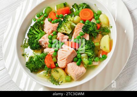 salmone, piselli, broccoli, patate, sedano, zuppa di carote e erbe verdi in una ciotola bianca su un tavolo di legno bianco, primo piano Foto Stock