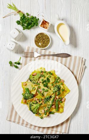 ravioli con verdure verdi, broccoli, piselli, asparagi, prosciutto tagliato a cubetti, prezzemolo e salsa cremosa su piatto bianco su tavolo di legno bianco con forchetta, ve Foto Stock