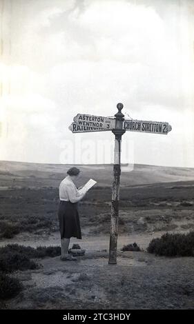 Anni '1950, storica, una signora che studia una mappa in piedi vicino a un cartello stradale nelle Shropshire Hills, Inghilterra, Regno Unito. Una freccia direzionale dice Church Stretton, una città mercato nello Shropshire, spesso chiamata "piccola Svizzera" in epoca vittoriana, per il suo paesaggio e la pittoresca ambientazione nelle Shropshire Hills. Foto Stock