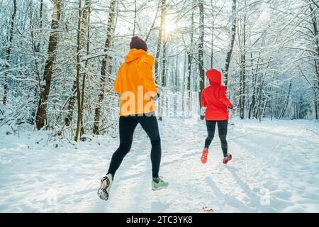 I giovani corridori di trail, uomo e donna, indossavano giacche antivento luminose che corrono nella pittoresca foresta innevata durante le soleggiate giornate gelate. Sport attivo p Foto Stock