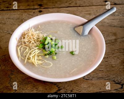 Immagine di cipolla fresca fresca fresca e fette di zenzero sul porridge di riso in un recipiente rosa chiaro con cucchiaio corto in metallo su un tavolo di legno, da vicino. Asiatico Foto Stock