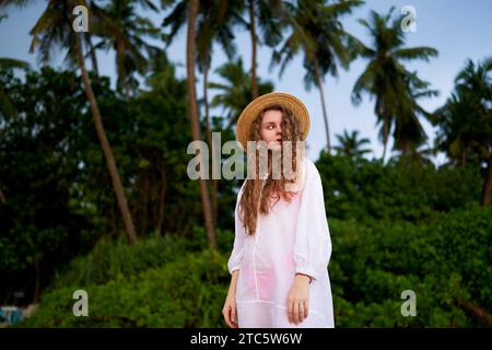 La giovane donna gode di una tranquilla fuga tropicale, vagando vicino alle palme da cocco con un elegante cappello da sole e un leggero vestito bianco, incarnando un'atmosfera bohémien estiva Foto Stock