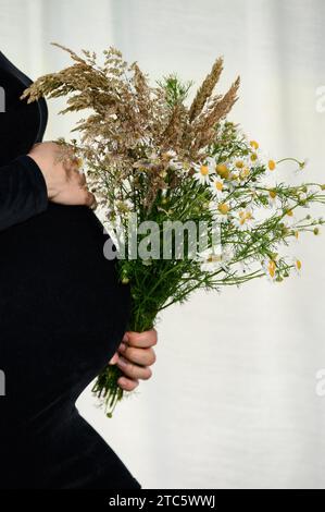 Una donna incinta sta abbracciando la pancia su uno sfondo bianco, indossa un vestito nero e tiene in mano un mazzo di fiori selvatici. Foto Stock