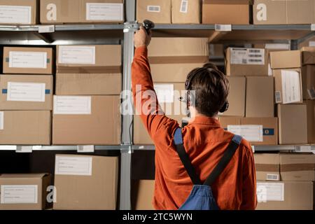 Operatore caucasico di magazzino che consegna merci, vestito con tute blu, si trova in piedi con la schiena girata e tira fuori diverse scatole di cartone Foto Stock