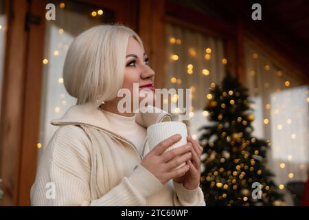 Bionda felice a casa a Natale, bevendo una bevanda calda dalla tazza bianca. Albero di Natale sullo sfondo. Festeggiamo Natale e Capodanno Foto Stock