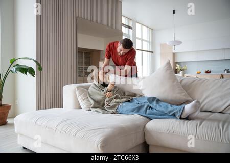 Marito premuroso e premuroso che controllava la temperatura della moglie malata sdraiata sul divano, toccando la fronte Foto Stock