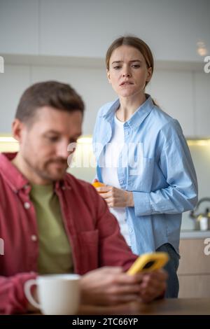 Donna che sbircia nello smartphone uomo in piedi dietro la schiena in cucina a casa. Foto Stock