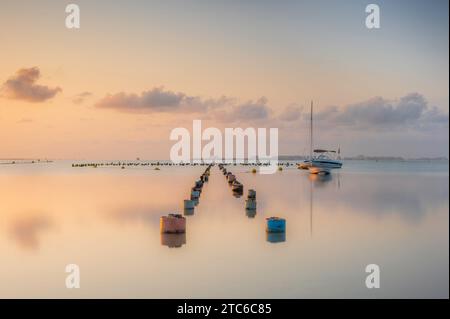 Posti da un molo in rovina, che si estendono in un mare calmo all'alba. Una piccola barca può essere vista all'orizzonte Foto Stock