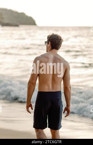 Schiena muscolosa, uomo in costume da bagno cammina sulla spiaggia dominicana al tramonto. Foto Stock