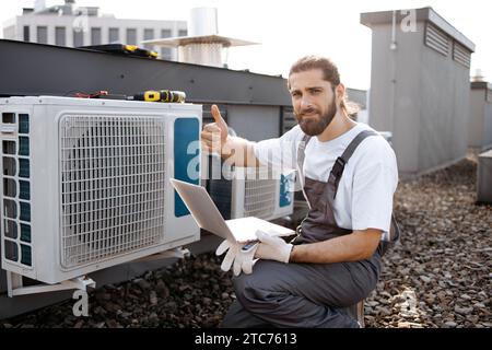 Artigiano che tiene il computer portatile e controlla il condizionatore sul tetto Foto Stock