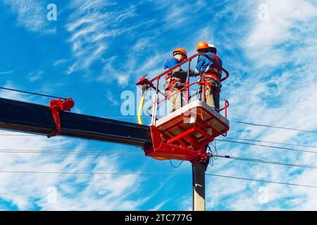 Due elettricisti dalla base della piattaforma aerea o della gru stanno riparando la lampada per illuminazione stradale. Elettricisti professionisti che indossano caschi, tute e assicurazioni in altezza. Vista dei lavoratori dal basso verso il cielo. Flusso di lavoro autentico. Foto Stock