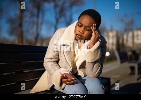 Ritratto all'aperto di una donna afroamericana preoccupata nelle giornate di sole. È seduta sulla panchina in strada e usa lo smartphone. Foto Stock