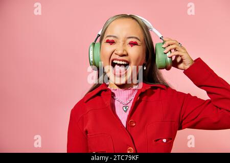 eccitata giovane donna asiatica che ascolta musica con cuffie wireless e urla su sfondo rosa Foto Stock