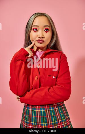 giovane donna asiatica pensiva con trucco occhi a forma di cuore che tocca la guancia e guarda lontano sul rosa Foto Stock