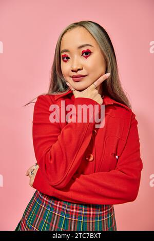 ritratto di una graziosa giovane donna asiatica con trucco oculare a forma di cuore che guarda la fotocamera su sfondo rosa Foto Stock