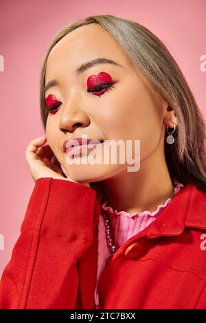 ritratto di una giovane donna asiatica pensiva con trucco oculare a forma di cuore che guarda lontano su sfondo rosa Foto Stock