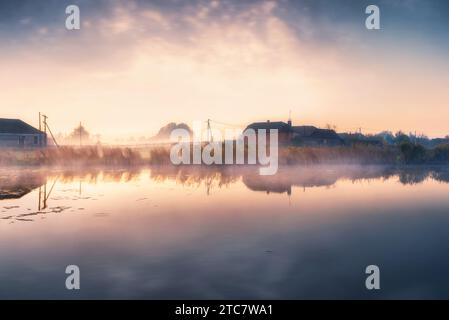 Splendida scena mattutina in un tranquillo villaggio con un lago nella nebbia all'alba. Campagna con case al sole all'alba. Foto Stock