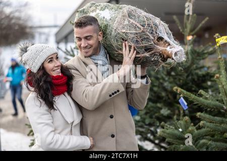 Un giovane che porta un albero di Natale avvolto, comprato al mercato insieme a sua moglie. La coppia felice ha comprato un albero di Natale. Foto Stock
