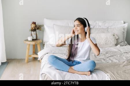 Una graziosa ragazza pre-adolescente ascolta musica tramite cuffie wireless, siediti a letto a casa goditi la canzone preferita in tutta tranquillità durante il weekend in camera da letto. Divertente, Leis Foto Stock