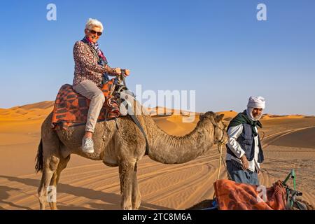 Cammellatore e turista occidentale cavalcando un cammello in una duna di sabbia di Erg Chebbi, deserto del Sahara vicino a Merzouga, Drâa-Tafilalet, Errachidia, Marocco Foto Stock