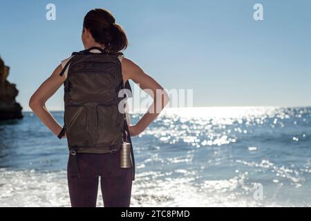 donna escursionista con zaino che guarda al mare sotto il sole Foto Stock