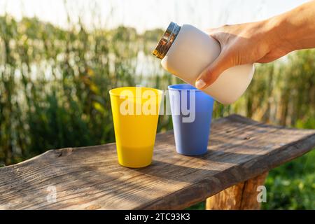 Una mano, versando il tè da un thermos bianco in una tazza blu su un tavolo di legno con accanto una tazza gialla. Lo sfondo è un lago con canne Foto Stock