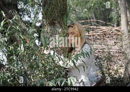 Bella donna bionda matura, capelli bagnati e bagnati, camicia bianca, occhi chiusi, abbracciare un tronco di salice, spazio per copiare Foto Stock