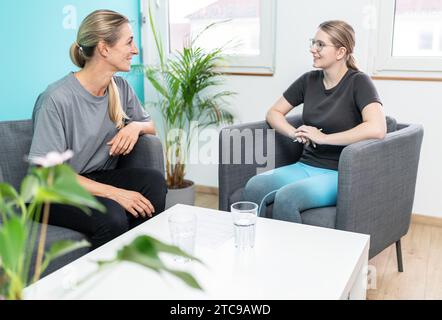 Due donne che conversano in una stanza luminosa, sedute su poltrone grigie Foto Stock