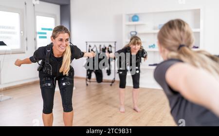 Due donne in tute da allenamento EMS che svolgono esercizi di equilibrio davanti a un trainer in palestra Foto Stock