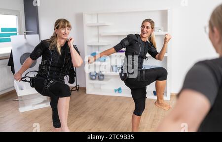 Due donne che si allenano in abiti di pronto soccorso in palestra sollevando la gamba e sorridendo con Trainer davanti a uno studio di pronto soccorso. Foto Stock