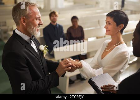 Vista laterale ritratto di coppie anziane sorridenti che si scambiano anelli durante la cerimonia di nozze in chiesa Foto Stock