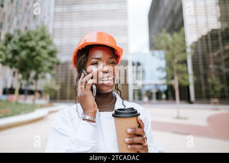 Felice donna nera con casco in piedi con una tazza di caffè e parlando sullo smartphone Foto Stock