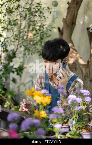 Giovane fiorista maschio asiatico felice e bello o proprietario di un piccolo giardino di fiori sta parlando al telefono con un fornitore che lavora nel suo giardino di fiori Foto Stock