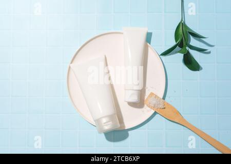 Due provette cosmetiche non etichettate vengono posizionate su una piastra rotonda in ceramica, un cucchiaio di sale e foglie verdi su uno sfondo blu pastello. Spazio raffinato per Foto Stock