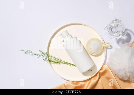 Una bottiglia bianca di gel doccia non etichettata è esposta su un piatto di ceramica con sale marino, un bicchiere d'acqua, una spugna doccia e un panno di seta su una b bianca Foto Stock