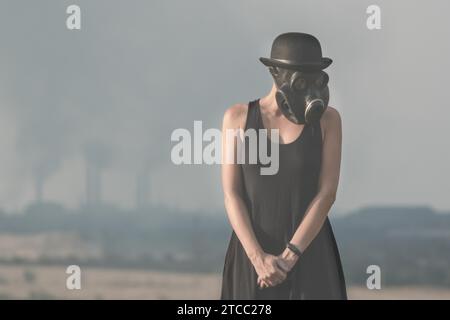 Giovane ragazza con un vestito nero e una maschera antigas sullo sfondo dei camini della fabbrica di fumo da vicino Foto Stock