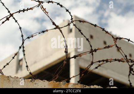 Vecchia recinzione in filo spinato arrugginito sullo sfondo di un edificio abbandonato e del cielo blu Foto Stock