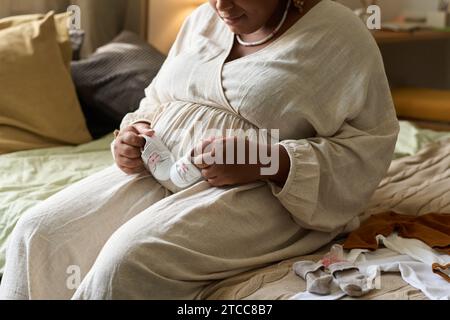 Mamma afroamericana seduta sul letto e tenendo i vestiti dei bambini, aspetta il suo bambino Foto Stock