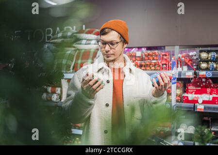 Un giovane con gli occhiali sta scegliendo regali natalizi per bambini nel supermercato, tenendo due giocattoli in mano e non può decidere su una scelta Foto Stock