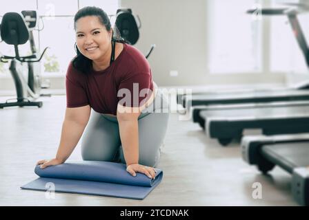 fat woman nello studio del club sportivo fitness con tappetino yoga. stuoia per pilates in rotolo femminile portrait plus happy smiling dopo la lezione di allenamento. Foto Stock