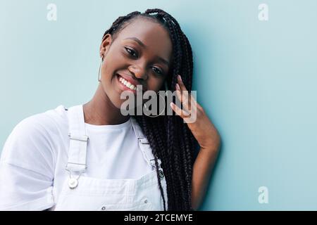 Ritratto ravvicinato di una giovane donna con trecce. Donna sorridente in abiti bianchi. Donna positiva che si appoggia a una parete blu e si regola le trecce mentre guarda Foto Stock